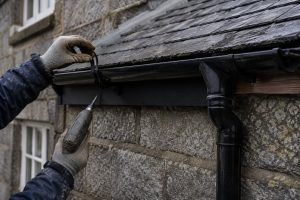 Black half-round gutter bracket being fitted to the fascia of a granite Aberdeen property, wet slate roof above