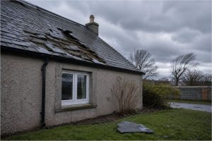 Slipped and missing slates on a harled Aberdeenshire cottage after a storm, broken slate on the grass below